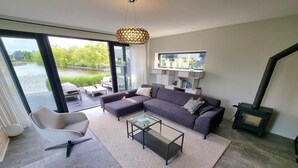 A modern living room with a sofa, armchair, and coffee table, large windows with a view of a water body, and a fireplace. - Waterfront Villa w/ Jetty (Balk)