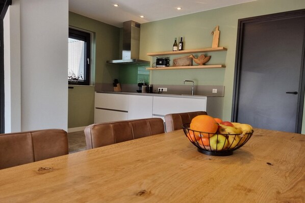 Une cuisine moderne équipée d’un four encastré, d’une cuisinière et d’un four à micro-ondes. On y trouve une table en bois, sur laquelle est posé un panier de fruits.
