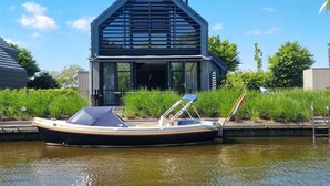 Una casa moderna con techo inclinado y ventanas grandes, un barco amarrado en la parte delantera y abundante vegetación alrededor.