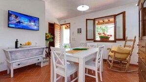 A dining area with a white table and chairs, a wooden rocking chair, a TV on the wall, and a window with a view of greenery. - Casa de' Fossi (Reggello)