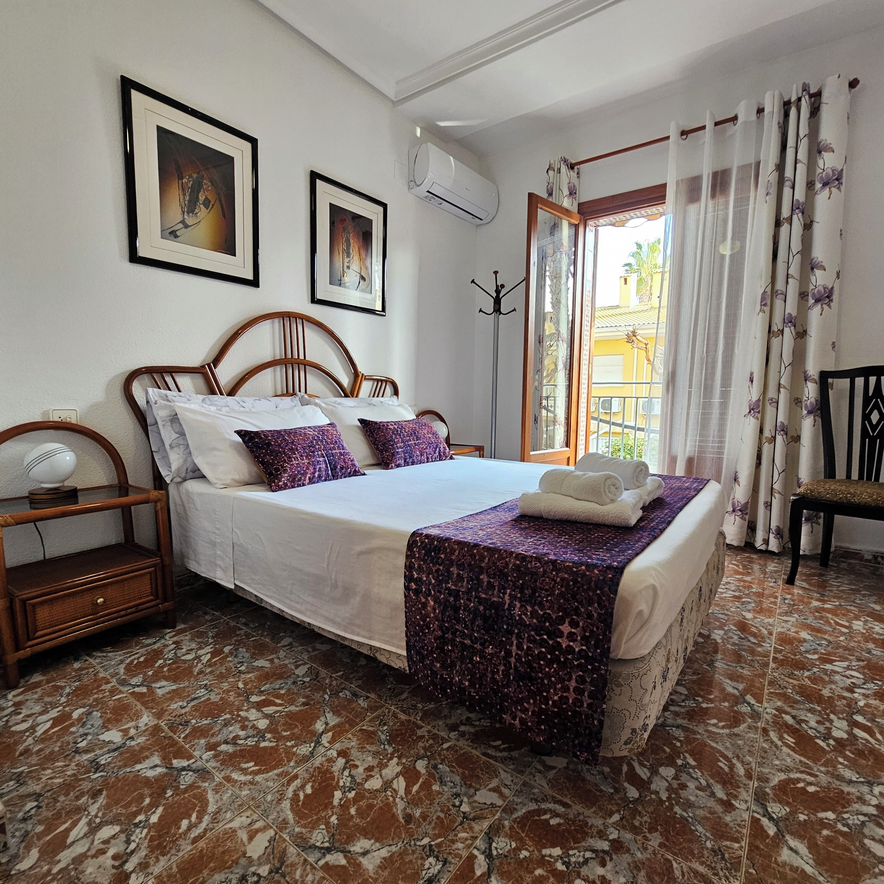 Un dormitorio ordenado con cama, mesita de noche, silla y ventana con cortinas.