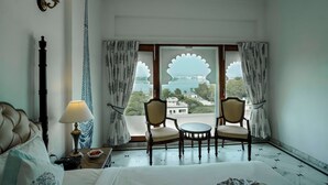 A hotel room with a bed, a nightstand, a lamp, two chairs, a table, and a large window with a view of the sea and buildings. - Burj Baneria - A Boutique Homestay (Udaipur)