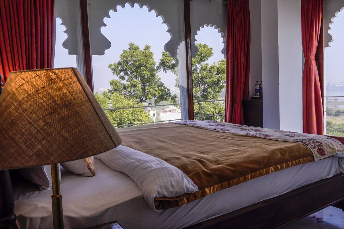 A bed with a patterned bedspread, a lamp, and a view of greenery through a window with decorative cutouts.