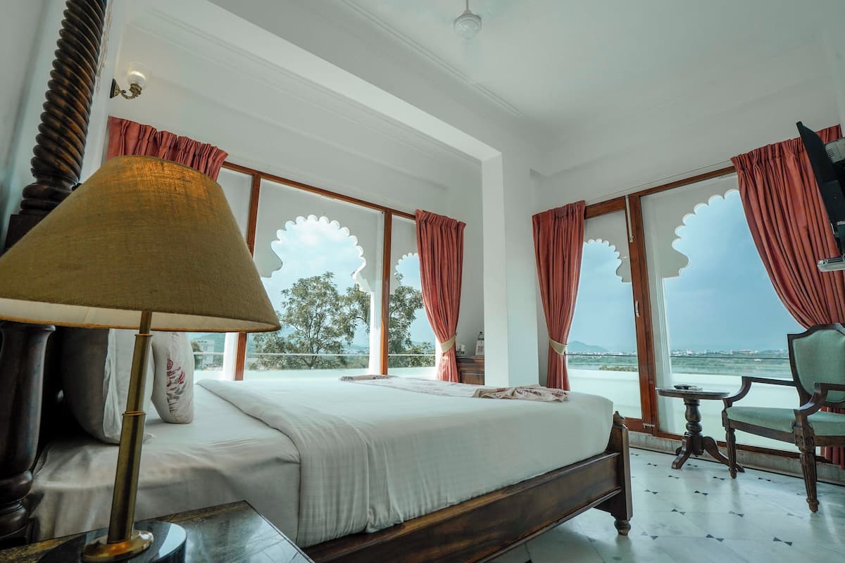 A hotel room with a bed, a table, and a chair. There are windows with curtains and a view of the outside.