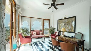 A room with a ceiling fan, a desk with a lamp, a sofa, and a view through a window with decorative glass panels. - Burj Baneria - A Boutique Homestay (Udaipur)