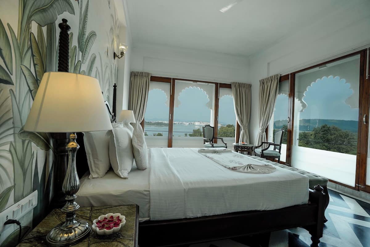 A hotel room with a bed, a lamp, a view of the sea, and a balcony with chairs.