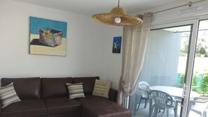 Un salon avec un canapé d’angle marron, un luminaire suspendu en fibres tressées et une porte en verre donnant sur une terrasse extérieure équipée d’une table et de chaises.
