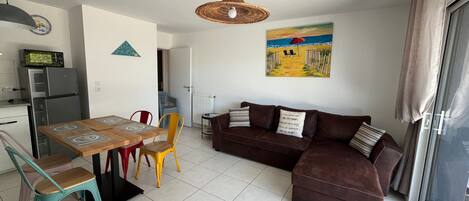 A living area with a brown sofa, a dining table with chairs, a kitchen with a refrigerator, and a painting on the wall.