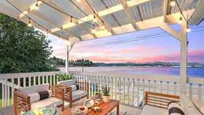 Un espace extérieur aménagé, abrité et doté d’un foyer, de mobilier en bois et offrant une vue sur un plan d’eau au coucher du soleil.