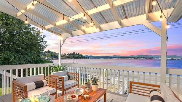 Un área de estar al aire libre techada, con una chimenea, muebles de madera y vista a un cuerpo de agua al atardecer.