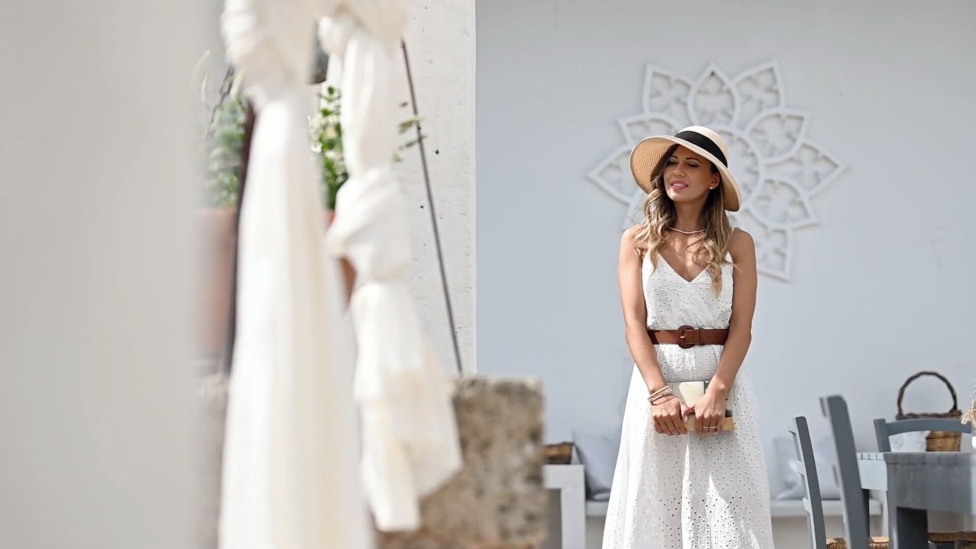 Une femme vêtue d’une robe blanche se tient devant un mur orné d’un motif décoratif.