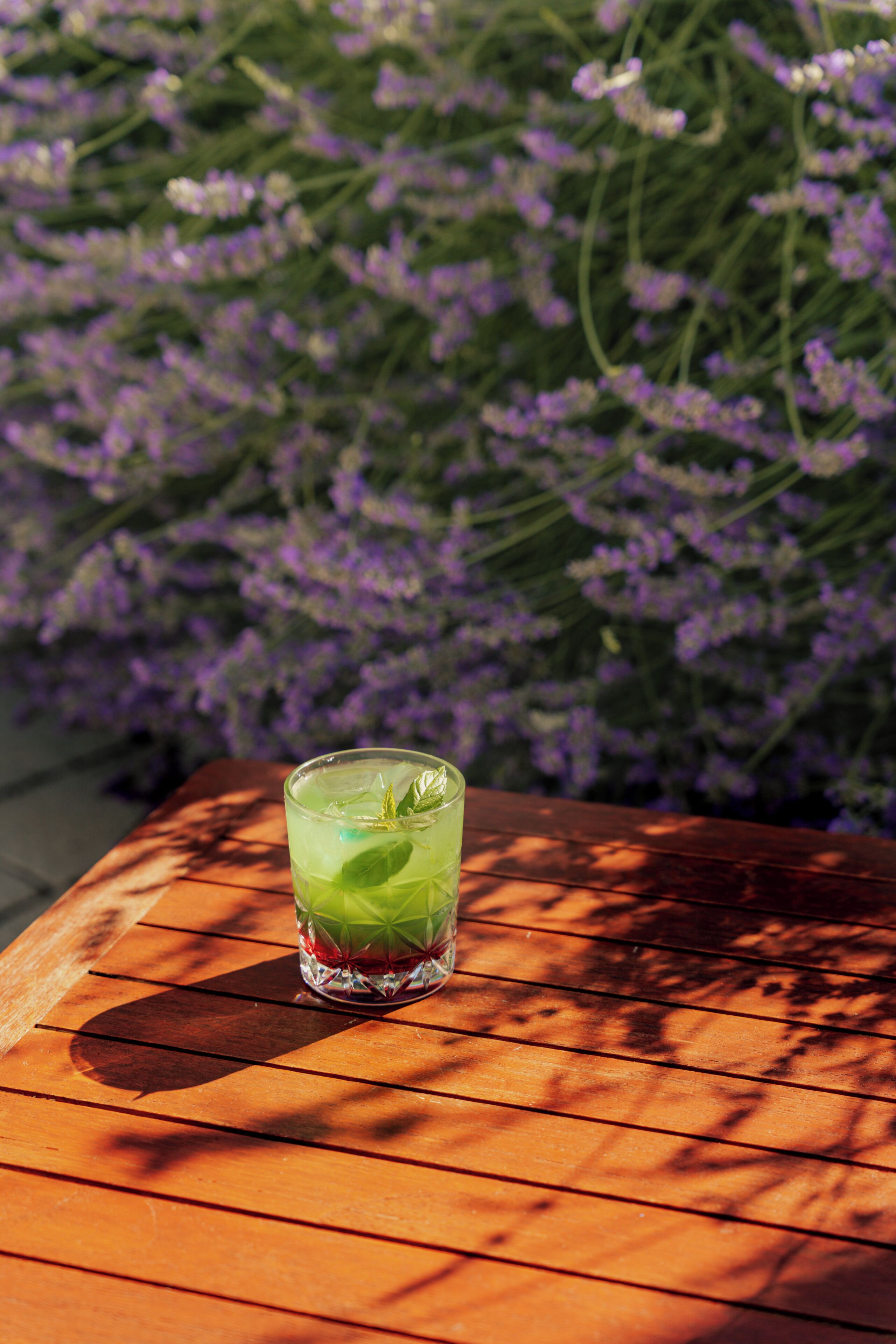 Un vaso de bebida verde sobre una mesa de madera con un fondo de flores moradas.
