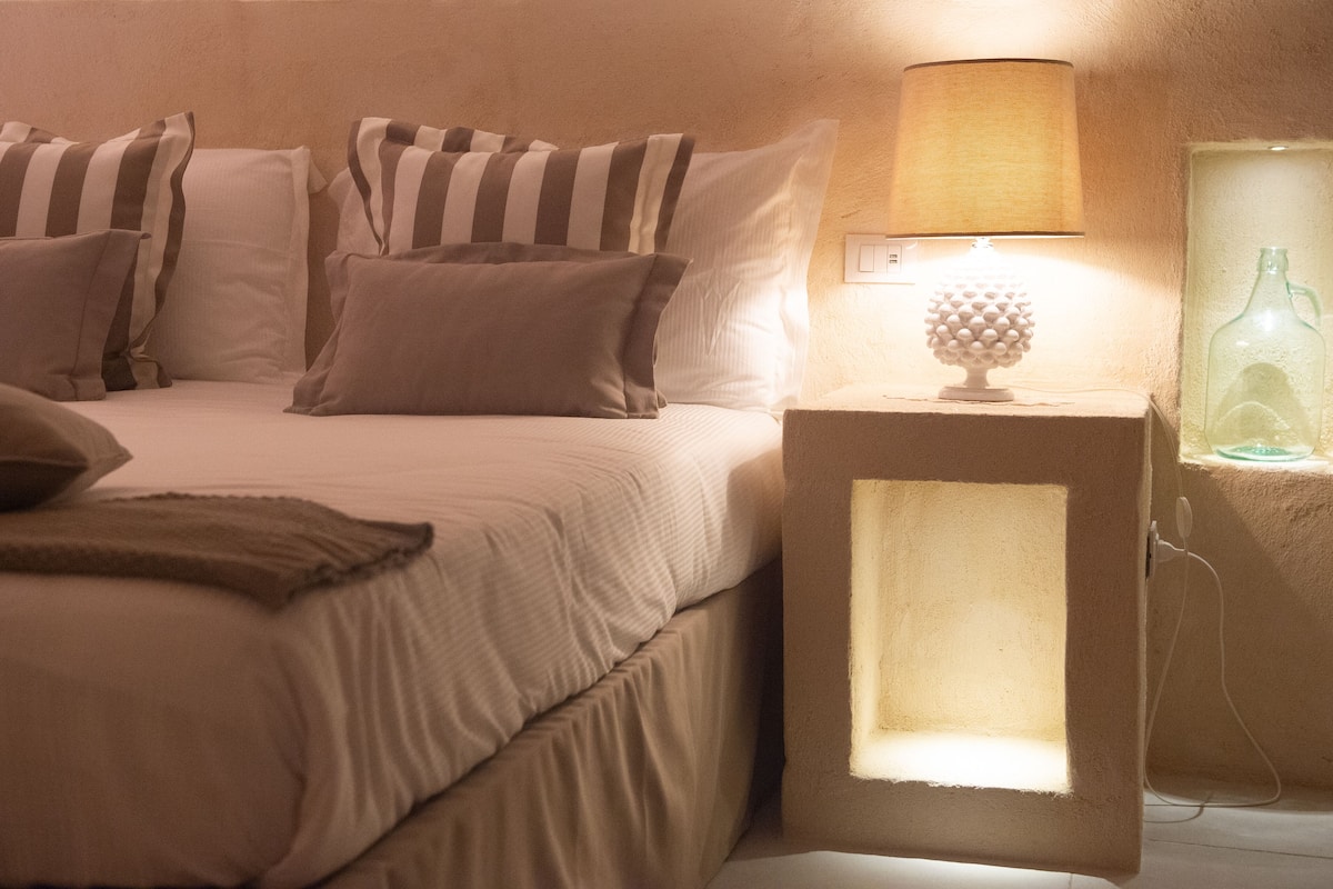 Une chambre à coucher avec un lit, des oreillers, une lampe de chevet et une table de chevet sur laquelle se trouve une bouteille.