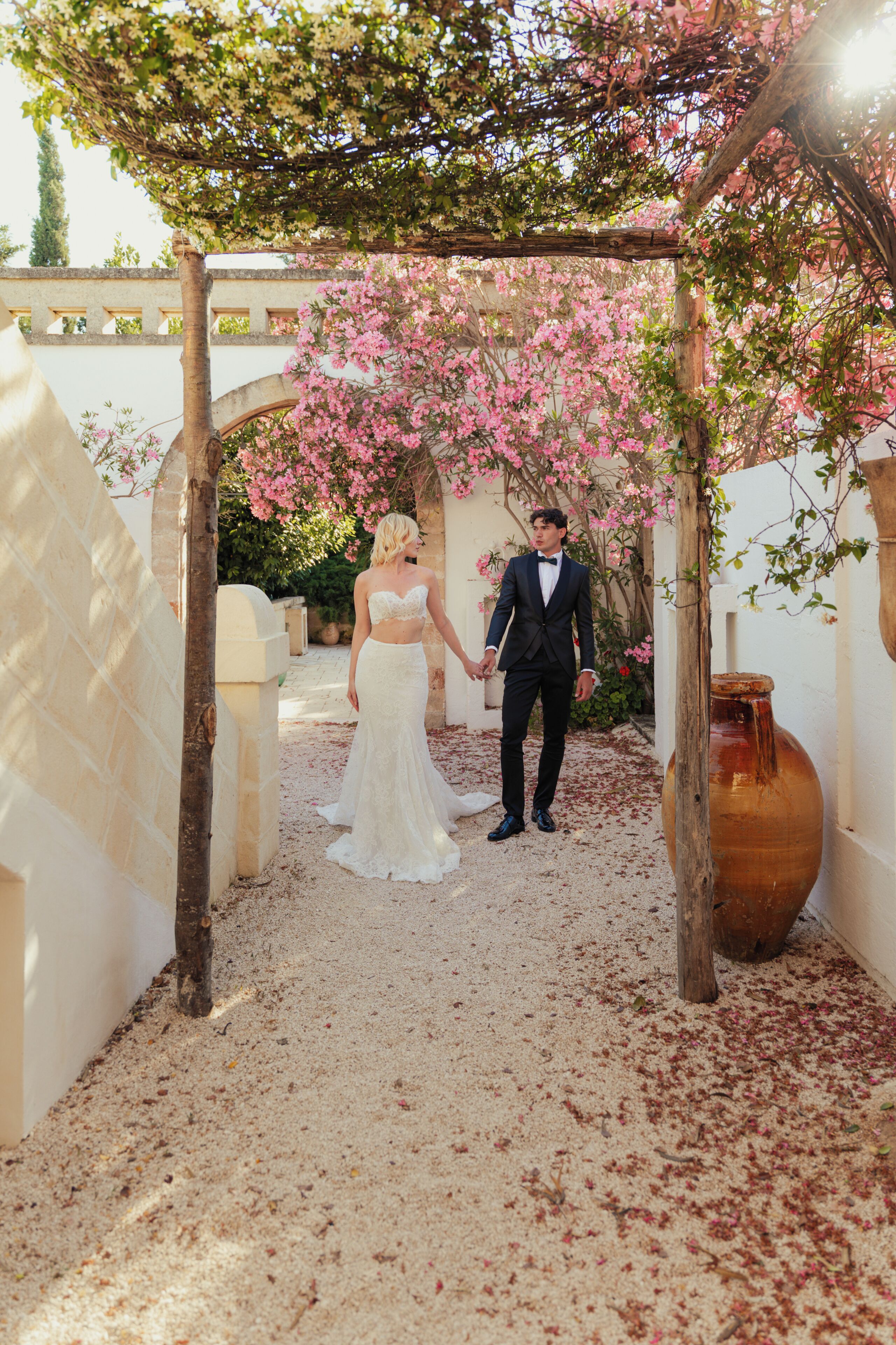 Una pareja bajo una pérgola con flores rosadas, un muro de piedra y un jarrón grande.