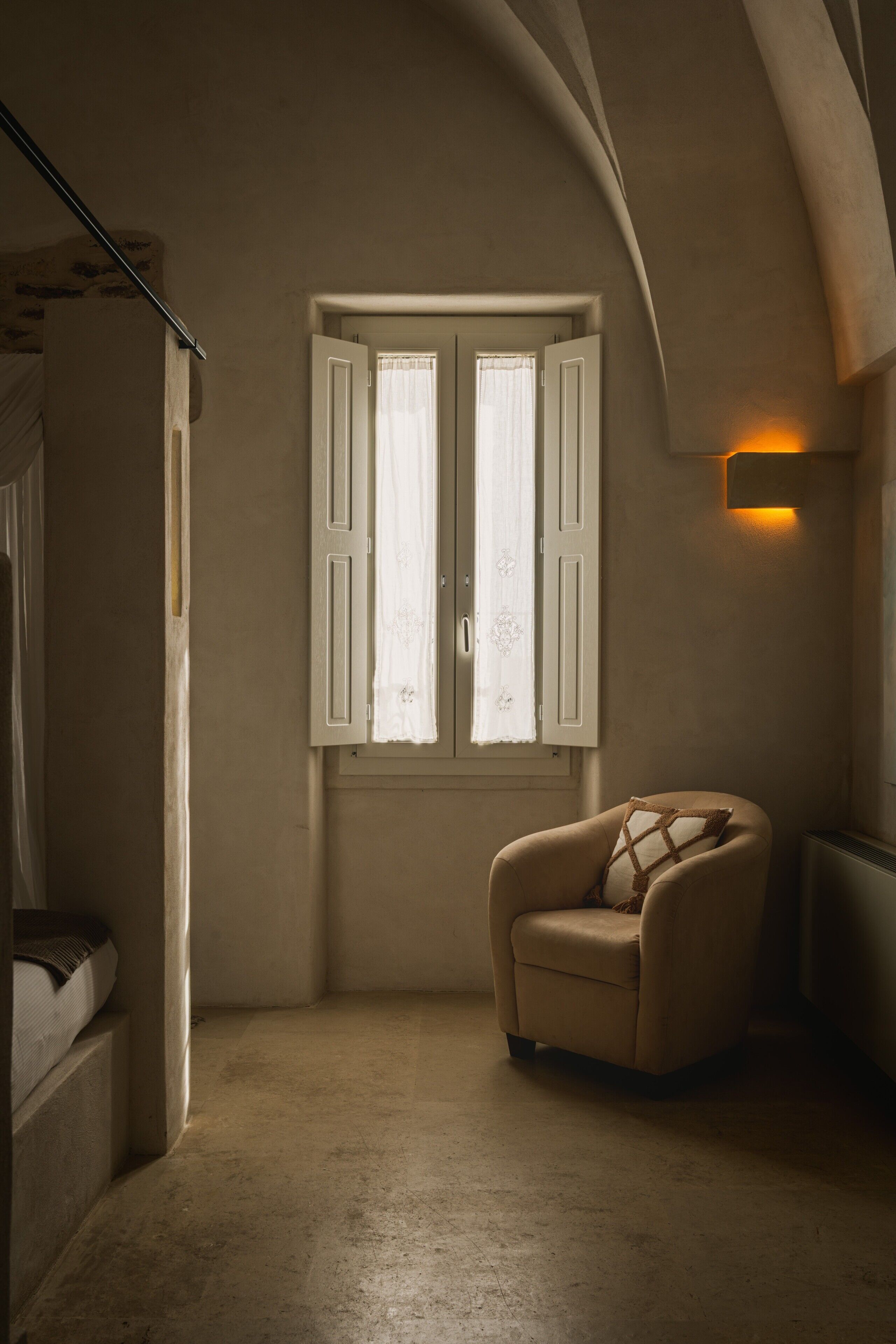 A room with a curved ceiling, a window with white shutters, a beige armchair with a cushion, and a wall-mounted light fixture.