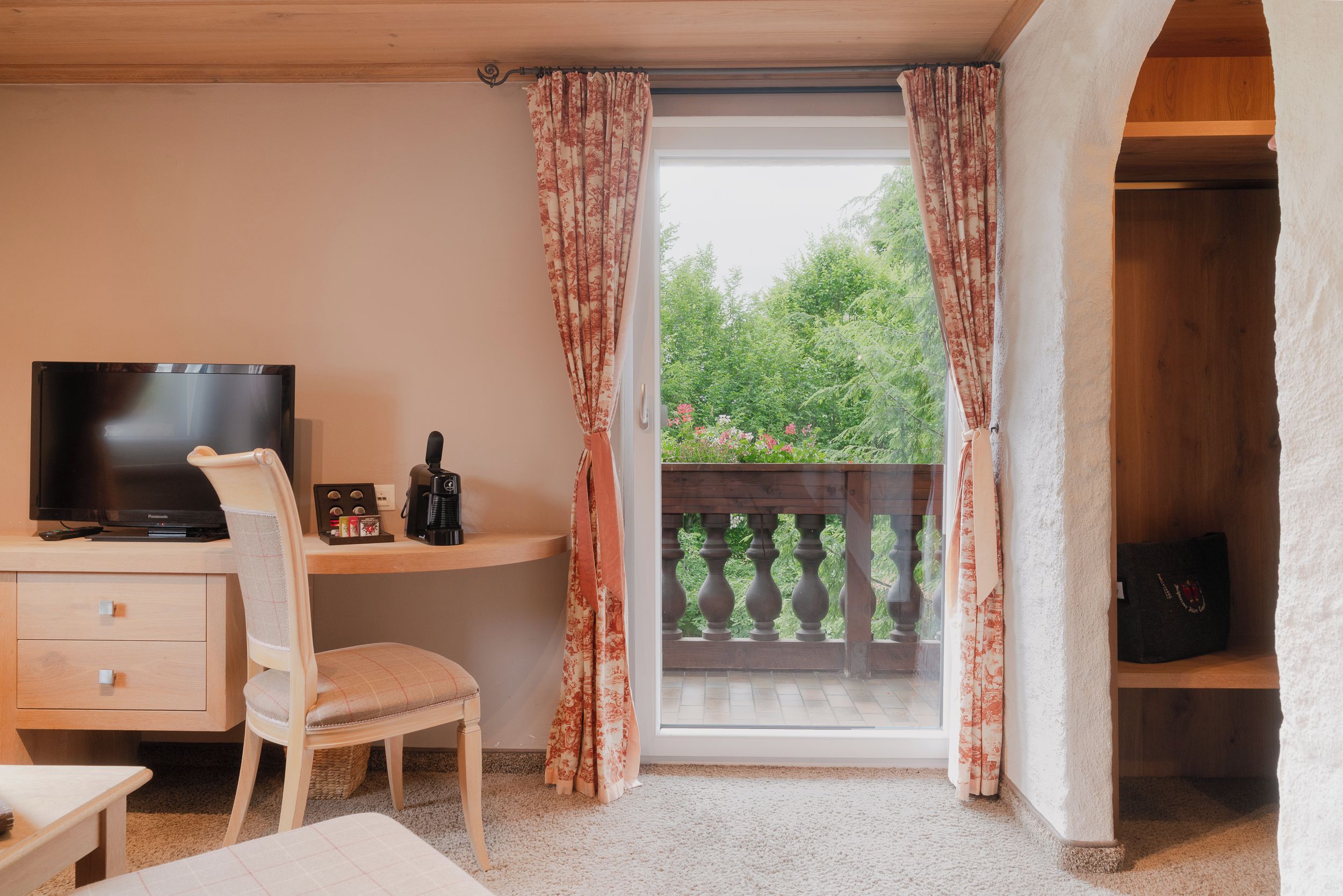 A room with a television, a desk, a chair, and a view of greenery through a window with curtains.
