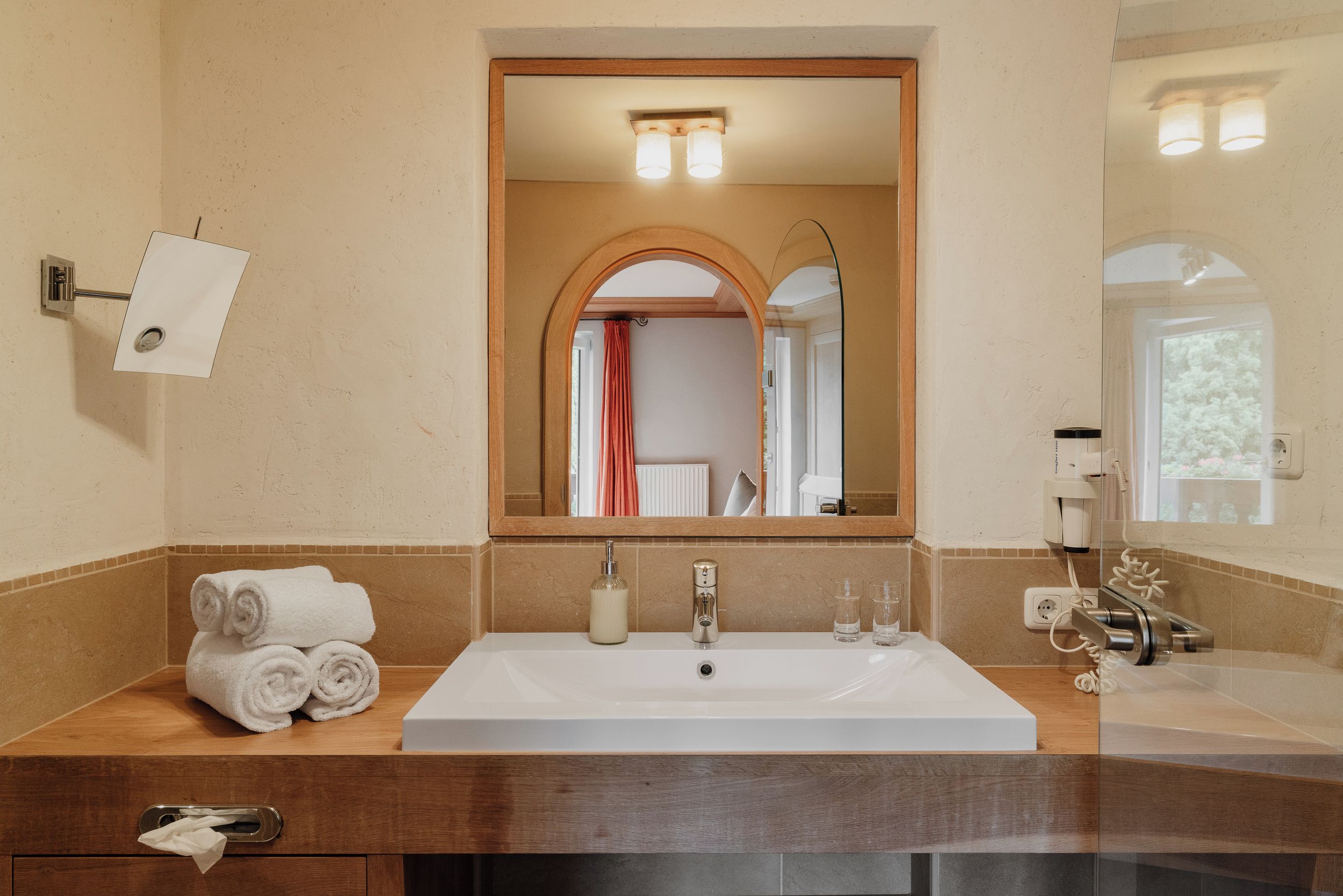 A bathroom with a large mirror, a sink, a towel dispenser, and a glass of water.
