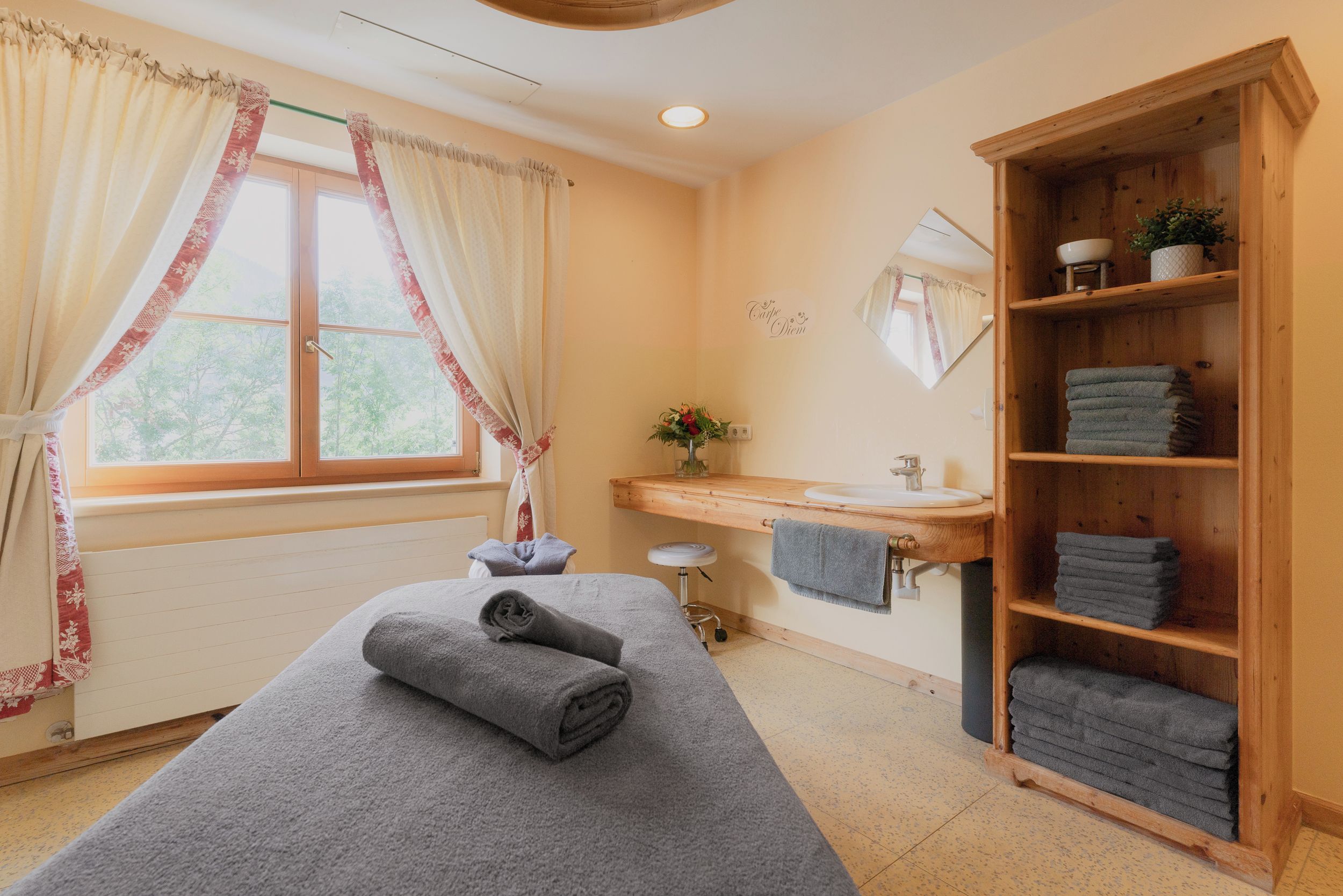 Quarto com mesa de massagem, prateleira de madeira com toalhas, janela com cortinas, pia e espelho.