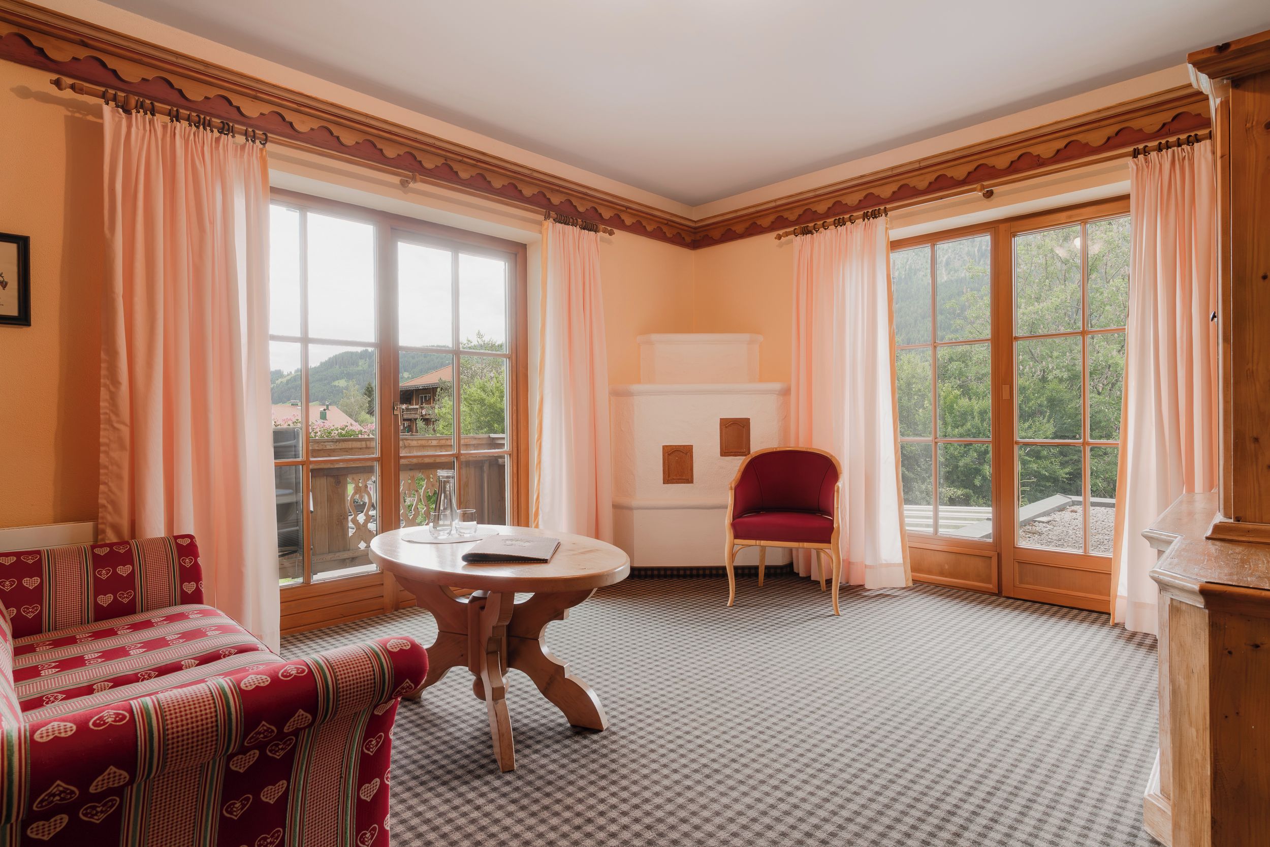 A room with a checkered floor, a round table, a red chair, and large windows with curtains.