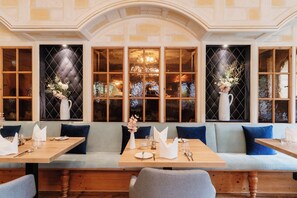 Un restaurante con una amplia zona de asientos tipo banco, varias mesas y grandes ventanales con jarrones decorativos.