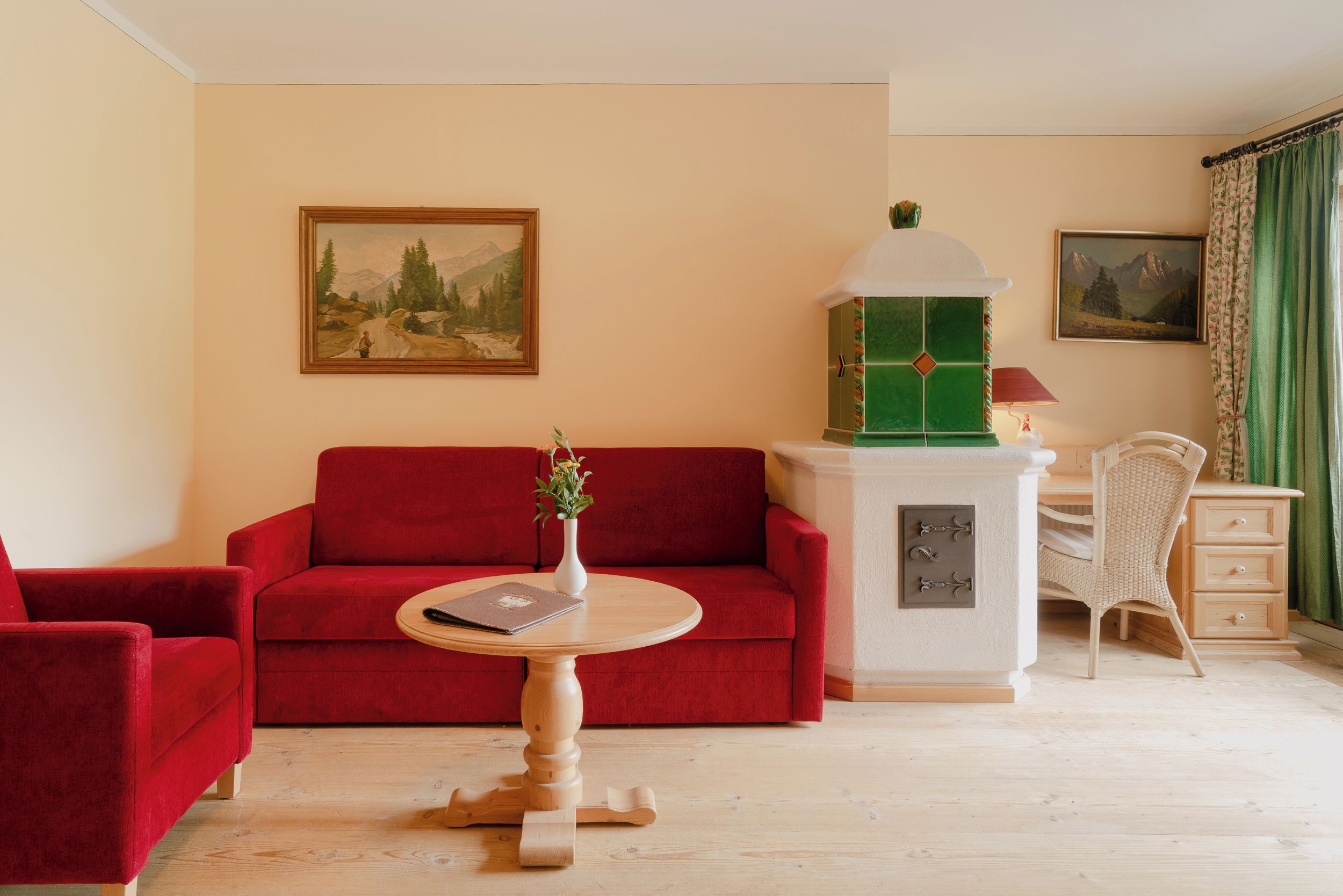 Ein Zimmer mit einem roten Sofa, einem kleinen runden Tisch mit einem Buch und einer Vase, einem grün-weißen Schrank, einem Korbstuhl und zwei gerahmten Bildern an der Wand.