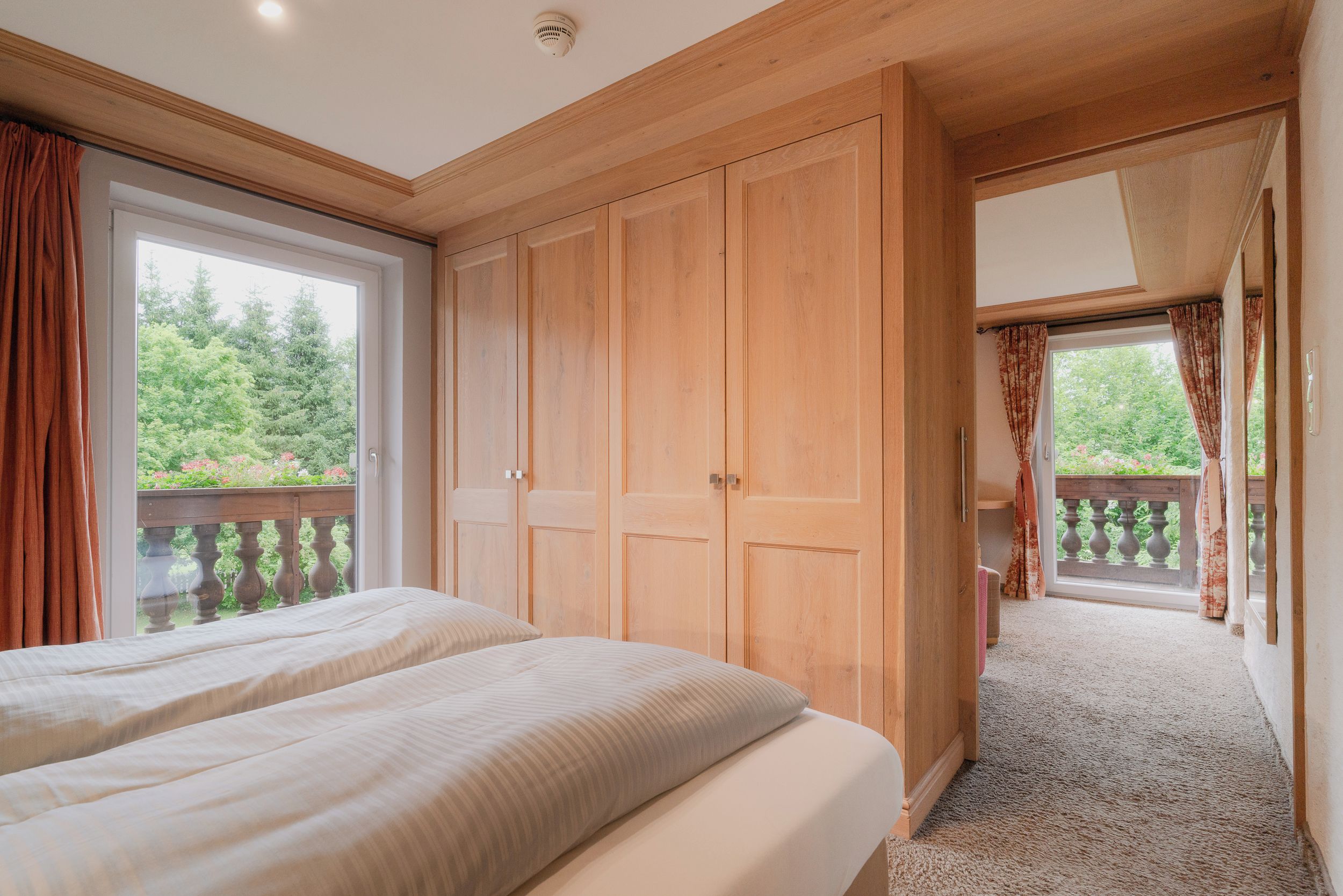Une chambre avec deux lits, une armoire en bois et un balcon donnant sur un espace verdoyant.