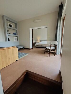 A hotel room with a bed, a desk, a chair, a small table, a shelf with decorative items, and a view of the hallway.