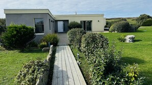 Ein modernes, zweistöckiges Haus mit einer Holzdiele, die zum Eingang führt, umgeben von gepflegten Gärten und blauem Himmel.