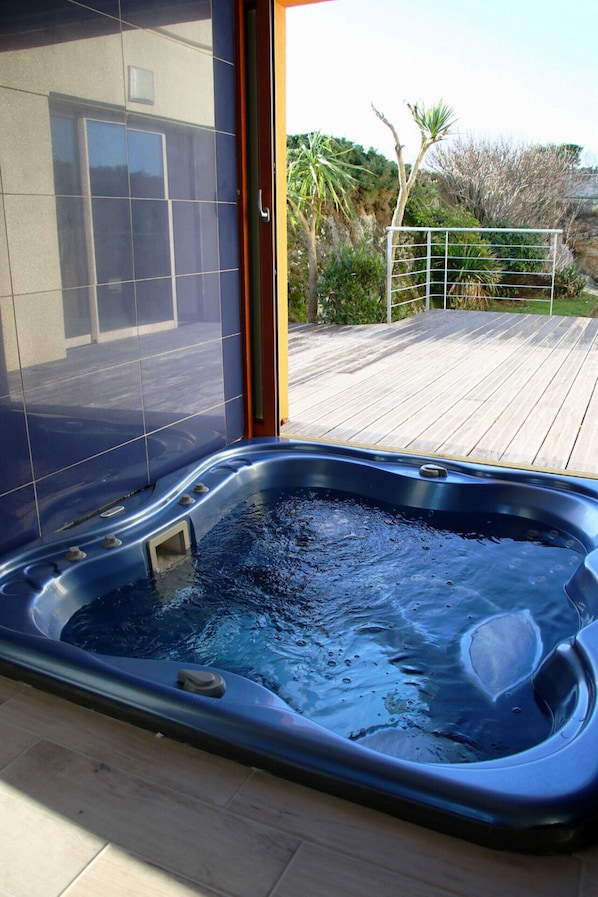 Un jacuzzi en una terraza de madera con vistas a un edificio y follaje.