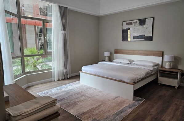 A modern bedroom with a large window, a bed, bedside tables, and a bench. - Al Faris Suite 2 (Manama)