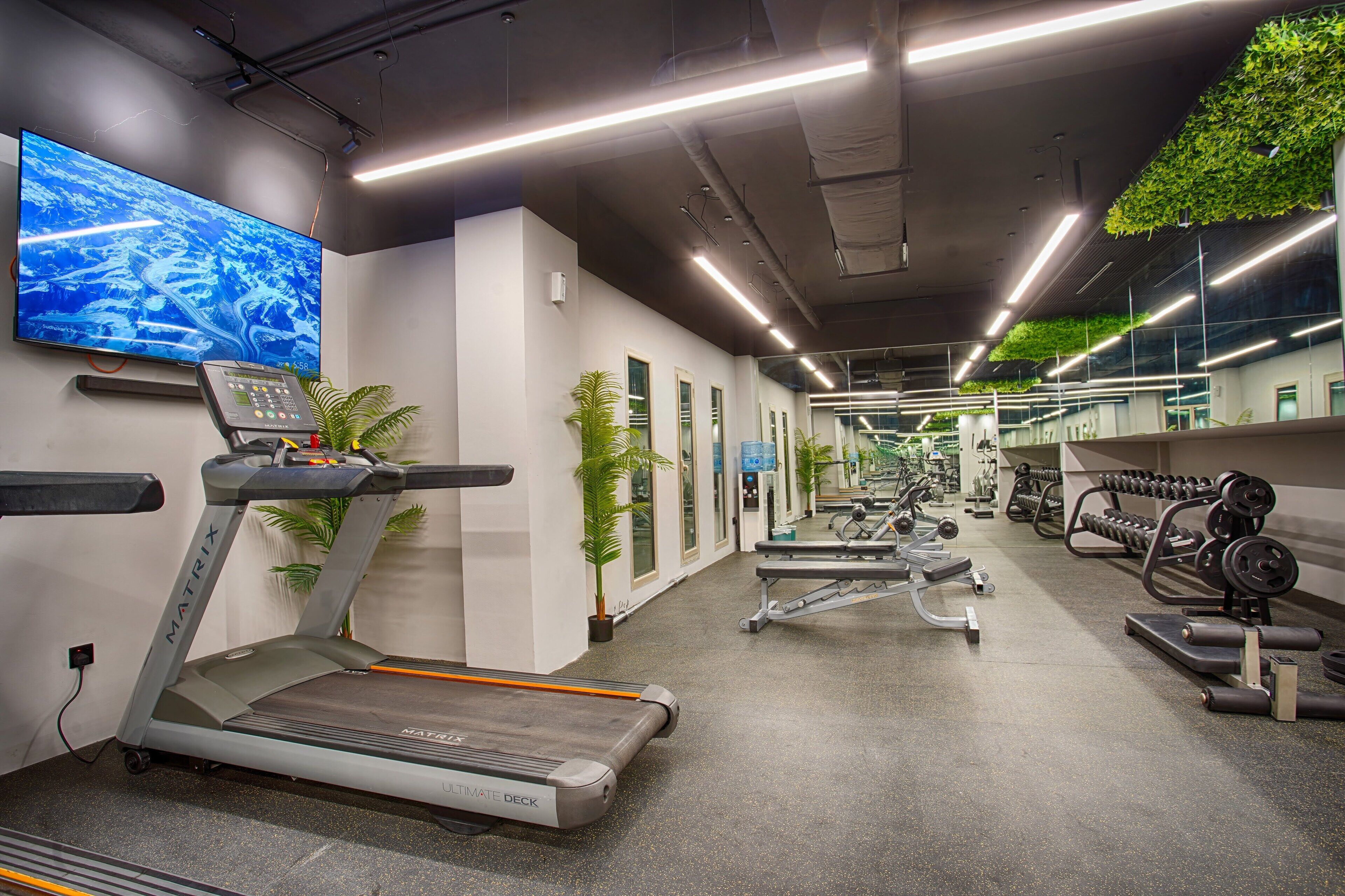 Une salle de sport moderne équipée de tapis de course, d’appareils de musculation et d’un grand écran de télévision diffusant des images prises par drone.
