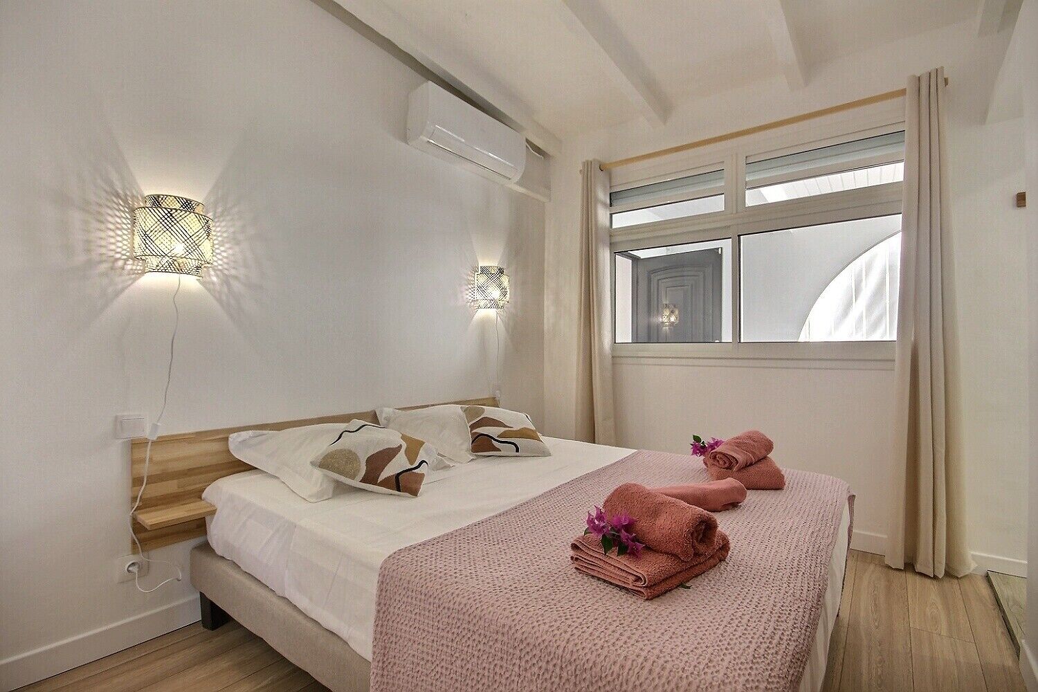 Una camera d'albergo con un letto, due luci fissate al muro, una finestra con tende e un pavimento in legno.