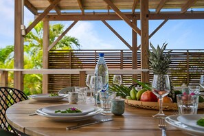 Un espace de restauration en plein air avec une table en bois dressée pour deux personnes, comprenant des assiettes, des couverts, des verres et une bouteille d’eau.