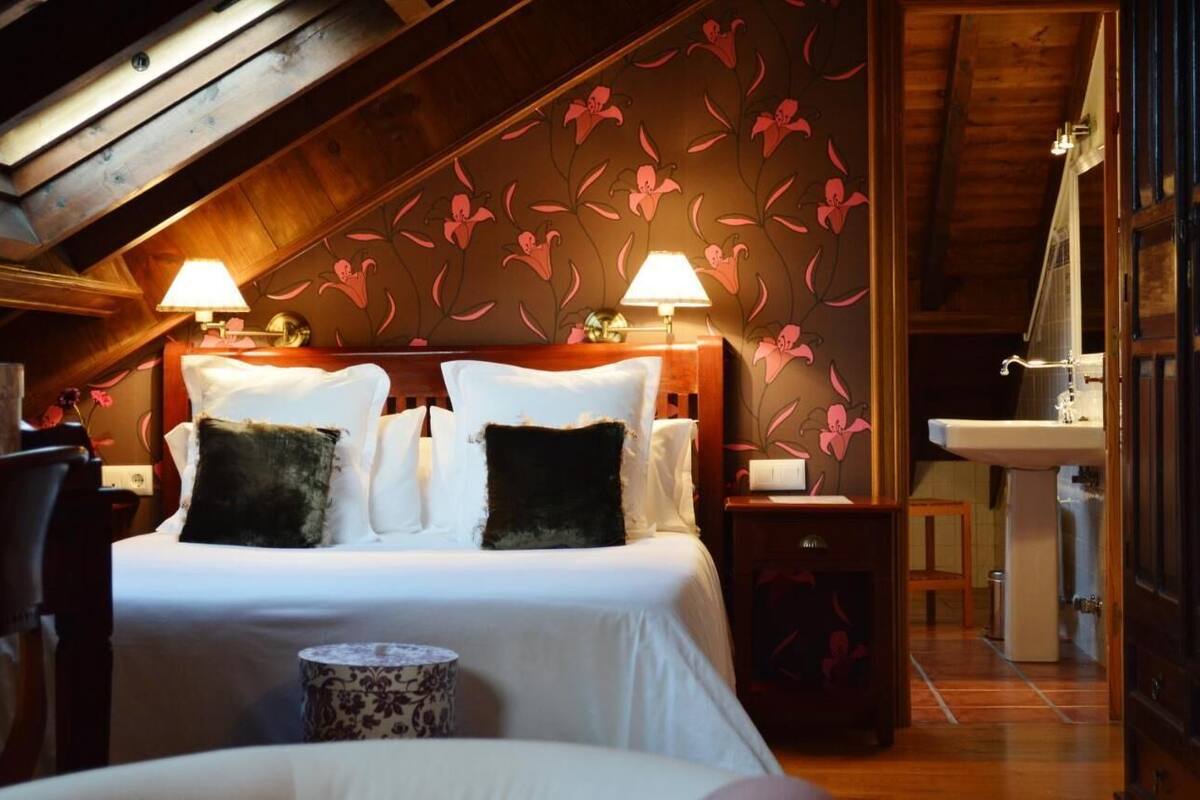 Une chambre avec un plafond en bois, un lit recouvert de draps blancs, deux lampes de chevet et un papier peint à motifs floraux.