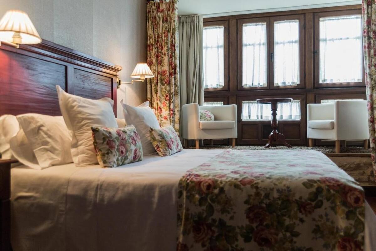 Une chambre d’hôtel avec un grand lit, deux fauteuils, une petite table et une vue sur l’extérieur grâce à de grandes fenêtres.