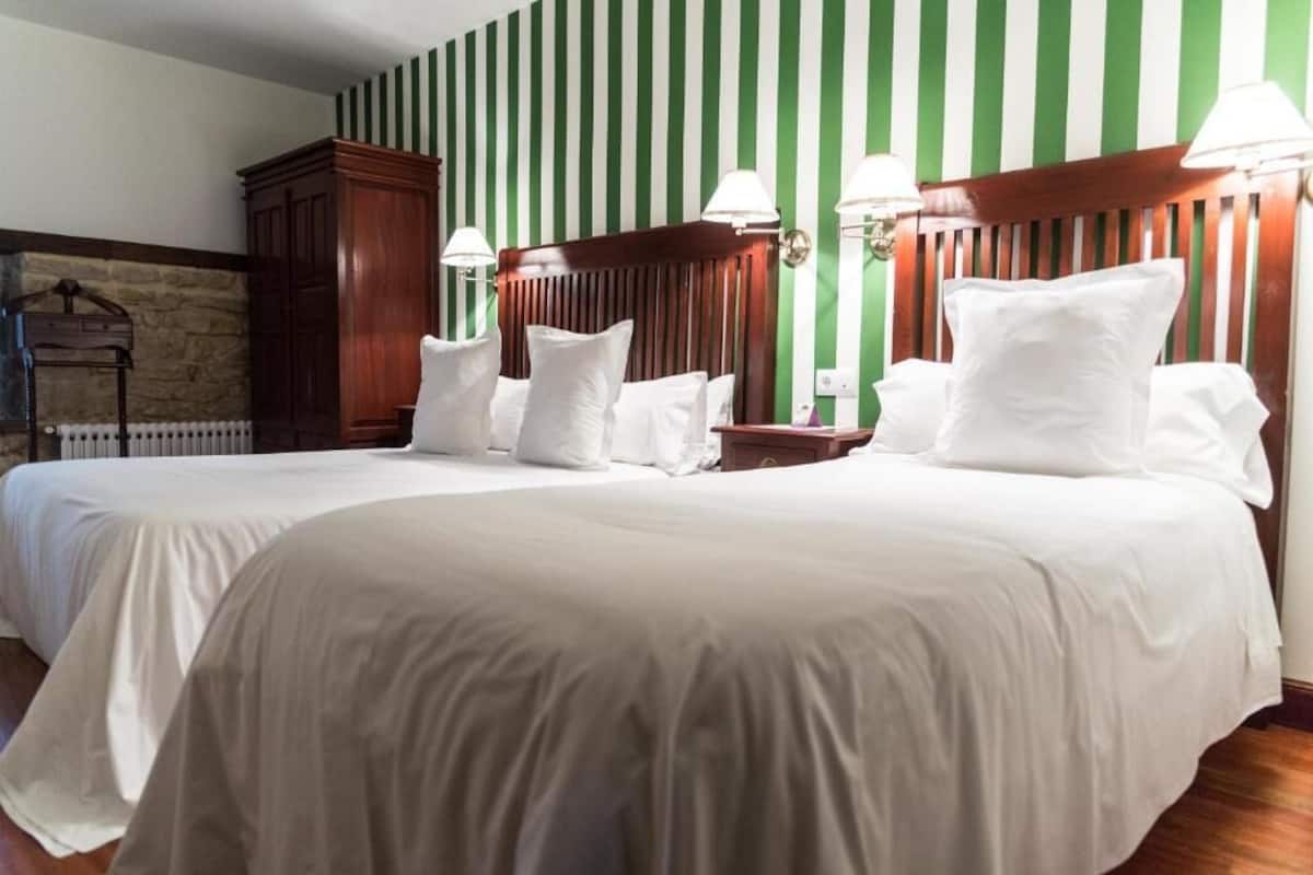 Une chambre d’hôtel avec deux lits, une tête de lit en bois, une cheminée en pierre et un papier peint à rayures vertes et blanches.