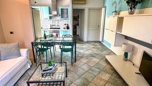 Un salon moderne avec un canapé, une table basse et un meuble pour la télévision. On aperçoit une cuisine à l’arrière-plan, avec un coin repas.