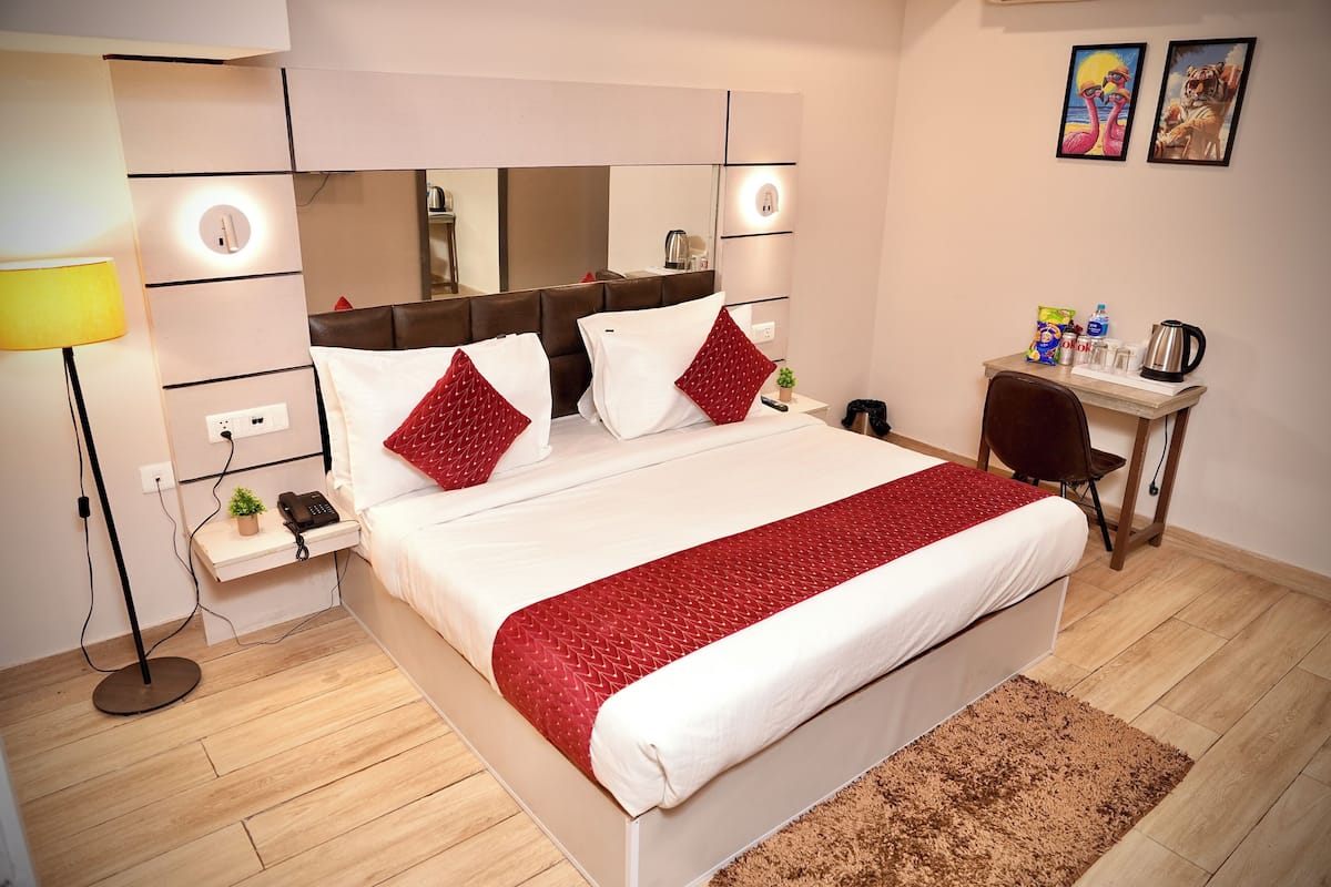 Habitación de hotel moderna con una cama grande, una mesita de noche con lámpara, un escritorio pequeño con silla y un espejo grande sobre la cama.