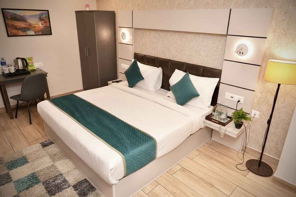 A hotel room with a large bed, a desk with a chair, a wardrobe, a bedside table with a lamp, and a small plant.