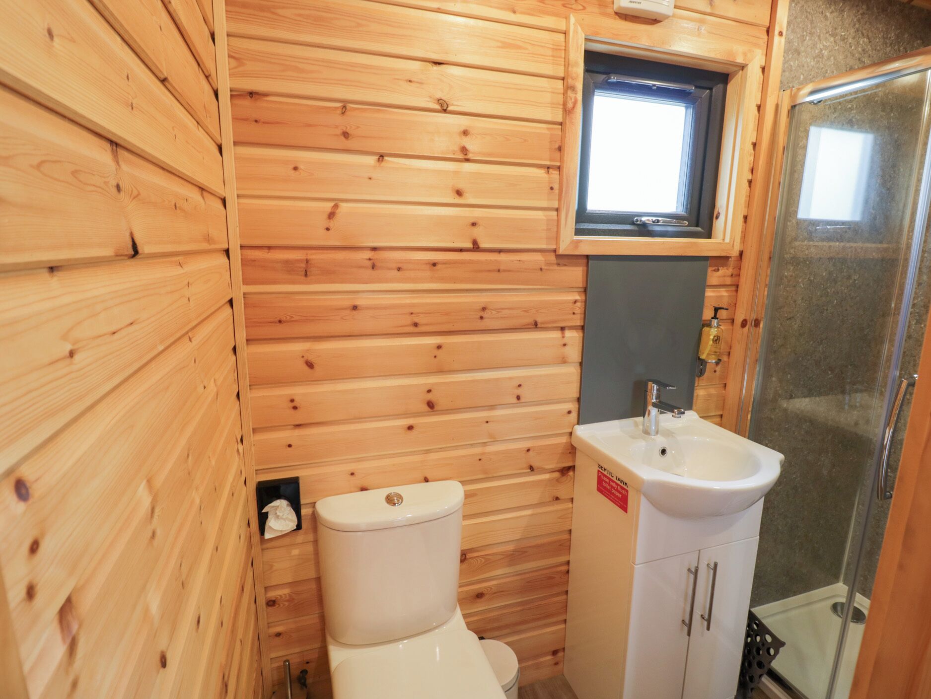 Une petite salle de bain en bois, équipée de toilettes, d’un lavabo et d’une petite fenêtre.