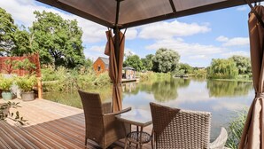 Eine Holzterrasse mit Korbmöbeln mit Blick auf einen ruhigen Fluss, umgeben von viel Grün und einer kleinen Holzkonstruktion.