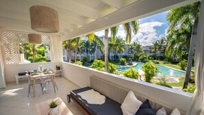 A balcony with a sofa, dining table, and chairs, overlooking a pool and palm trees. - Family Dominicus Apartment - Bayahibe (Bayahibe)