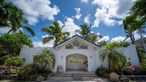A white building with a peaked roof and a central arched entrance, surrounded by palm trees and flowering plants. - Family Dominicus Apartment - Bayahibe (Bayahibe)