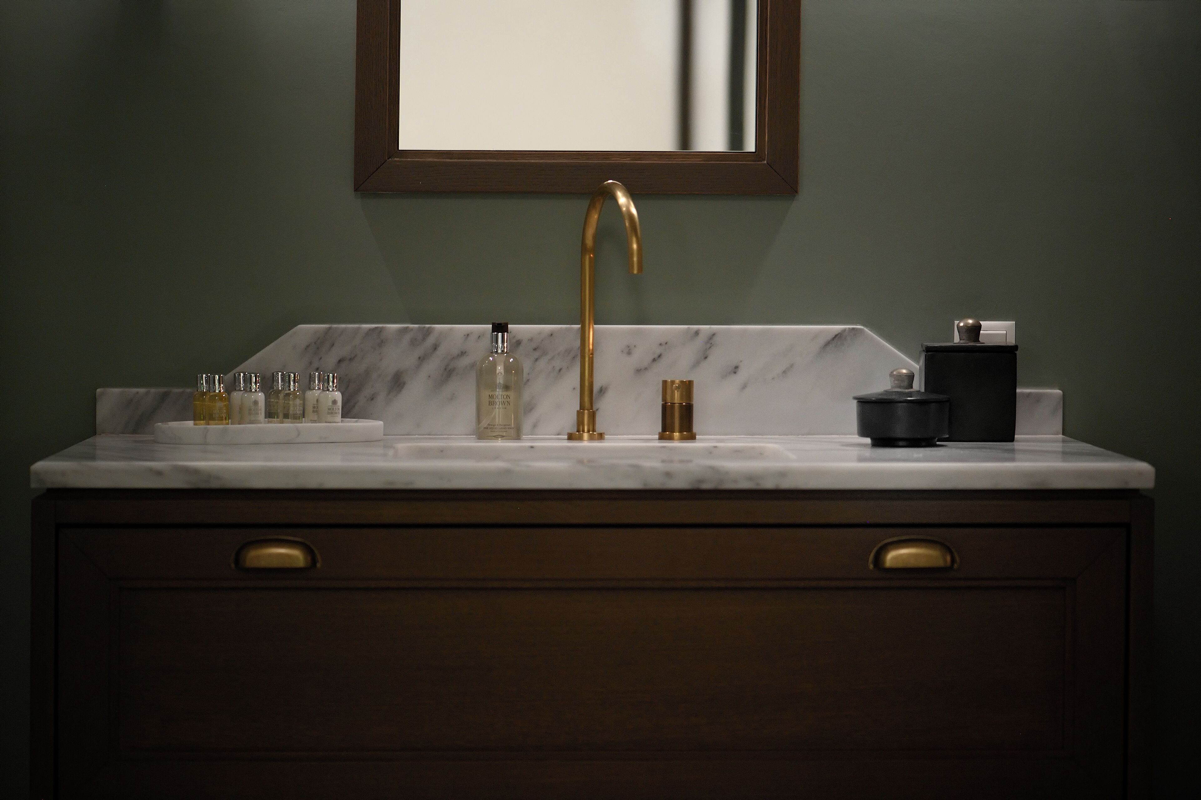 A bathroom with a marble sink, gold faucet, and a mirror above it. There are toiletries on the sink and a black container on the counter.