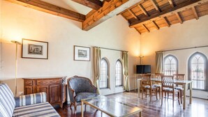 Un salón espacioso con vigas de madera, un sofá, una mesa de centro de vidrio, un televisor y un comedor con sillas y cortinas.