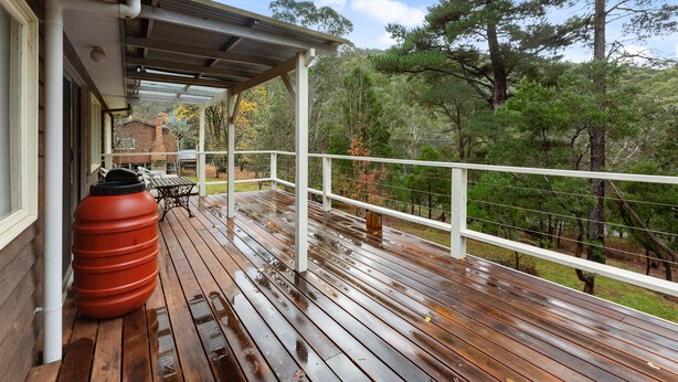 A wooden deck with a covered area, a large red water tank, and a view of trees and a brick building. - Club Snowflake - Base of Buller (Sawmill Settlement)