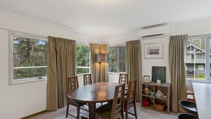 A dining area with a round wooden table and chairs, a small shelf with baskets, and a view of a house through large windows. - Club Snowflake - Base of Buller (Sawmill Settlement)