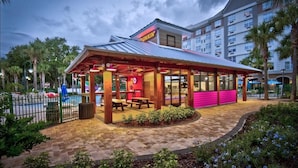 Un établissement DUNKIN' Donuts doté d’une terrasse couverte, de places assises en plein air et d’une piscine en arrière-plan.
