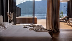 Een slaapkamer met een groot bed, uitzicht op een meer en een houten terras met een stoel.