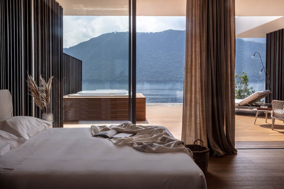 Een slaapkamer met een groot bed, uitzicht op een meer en een houten terras met een stoel.